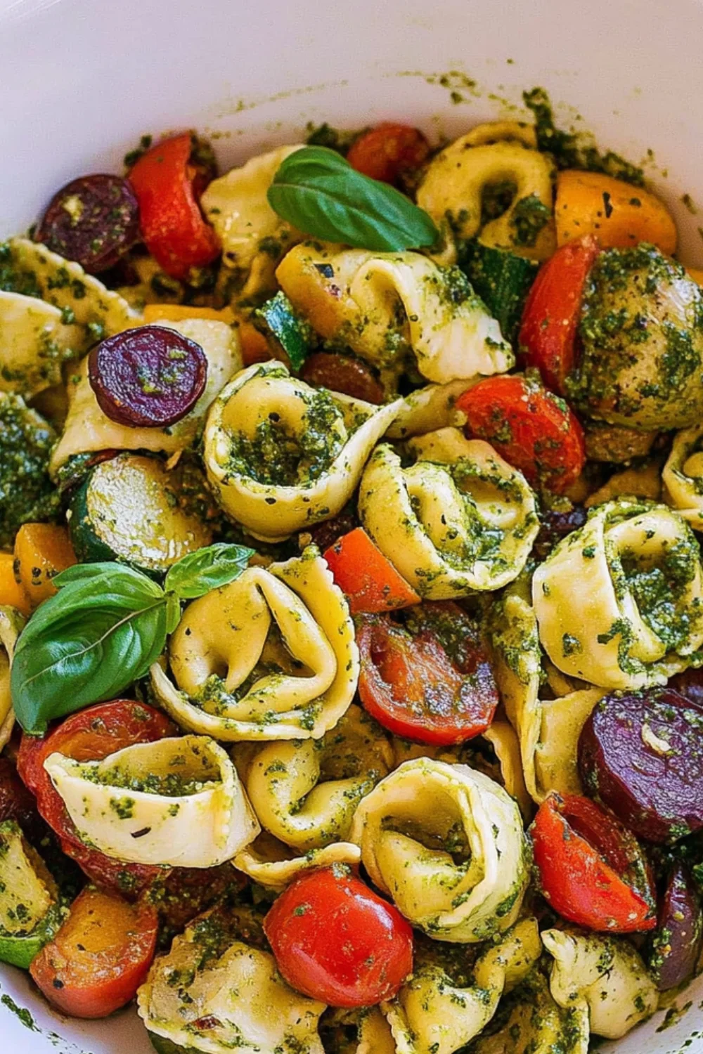 A close-up of Tortellini with Pesto and Roasted Veggies, featuring vibrant tortellini mixed with cherry tomatoes, roasted carrots, zucchini, and a generous drizzle of green pesto sauce, topped with fresh basil.
