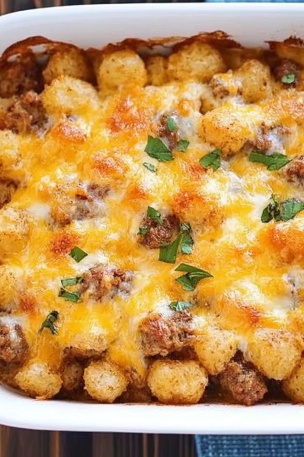 A delicious Tater Tot Sausage Breakfast Casserole with melted cheese, ground sausage, and crispy tater tots, garnished with fresh herbs in a white casserole dish.